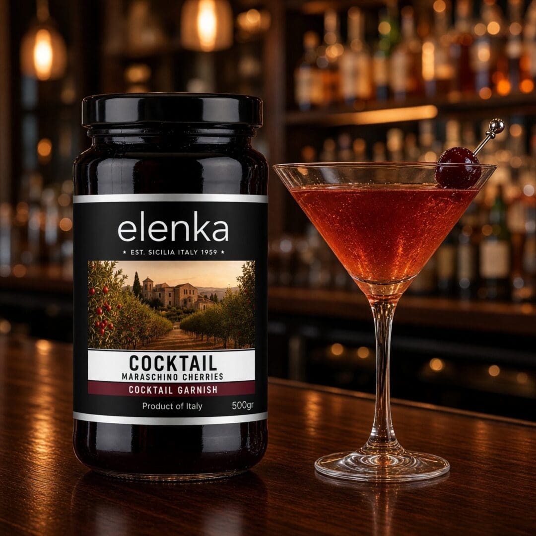 A jar of Elenka maraschino cherries is next to a cocktail glass garnished with a cherry, set on a polished bar with shelves of liquor bottles in the background.