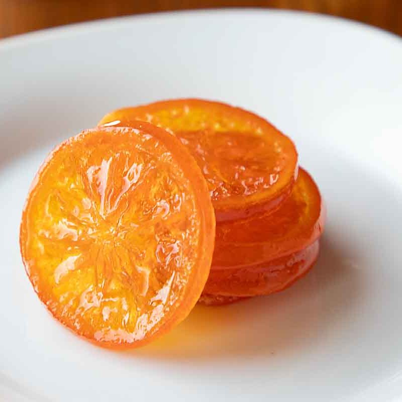 Slices of candied orange, reminiscent of classic Italian products, arranged on a white plate and glistening under the light, with a wooden table background.