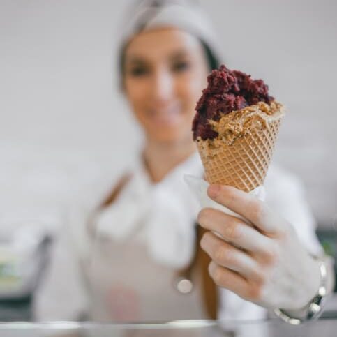 A person in a uniform and cap proudly presents a waffle cone brimming with purple and brown ice cream made from authentic Italian products. The background remains blurred, drawing all attention to the tempting treat.
