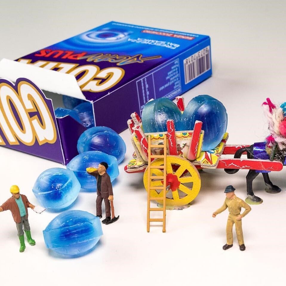 Miniature figures unload giant blue candy balls from a box labeled "GOLA Xtreme Plus," known for its Italian products, into a colorful cart pulled by a toy horse. Two figures stand nearby, and one is on a ladder, all arranged on a white surface.